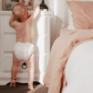 baby with white cloth nappy reaching up for toy ontop of drawers